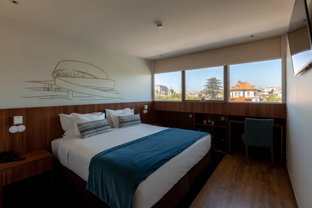 Modern hotel room with a large bed and a city view through wide windows.