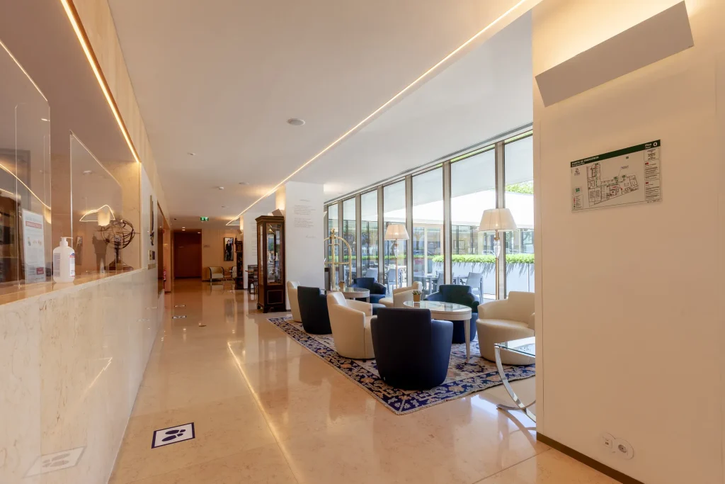 Hotel lobby with chairs, tables, lamps, and marble floor, bordered by large windows.