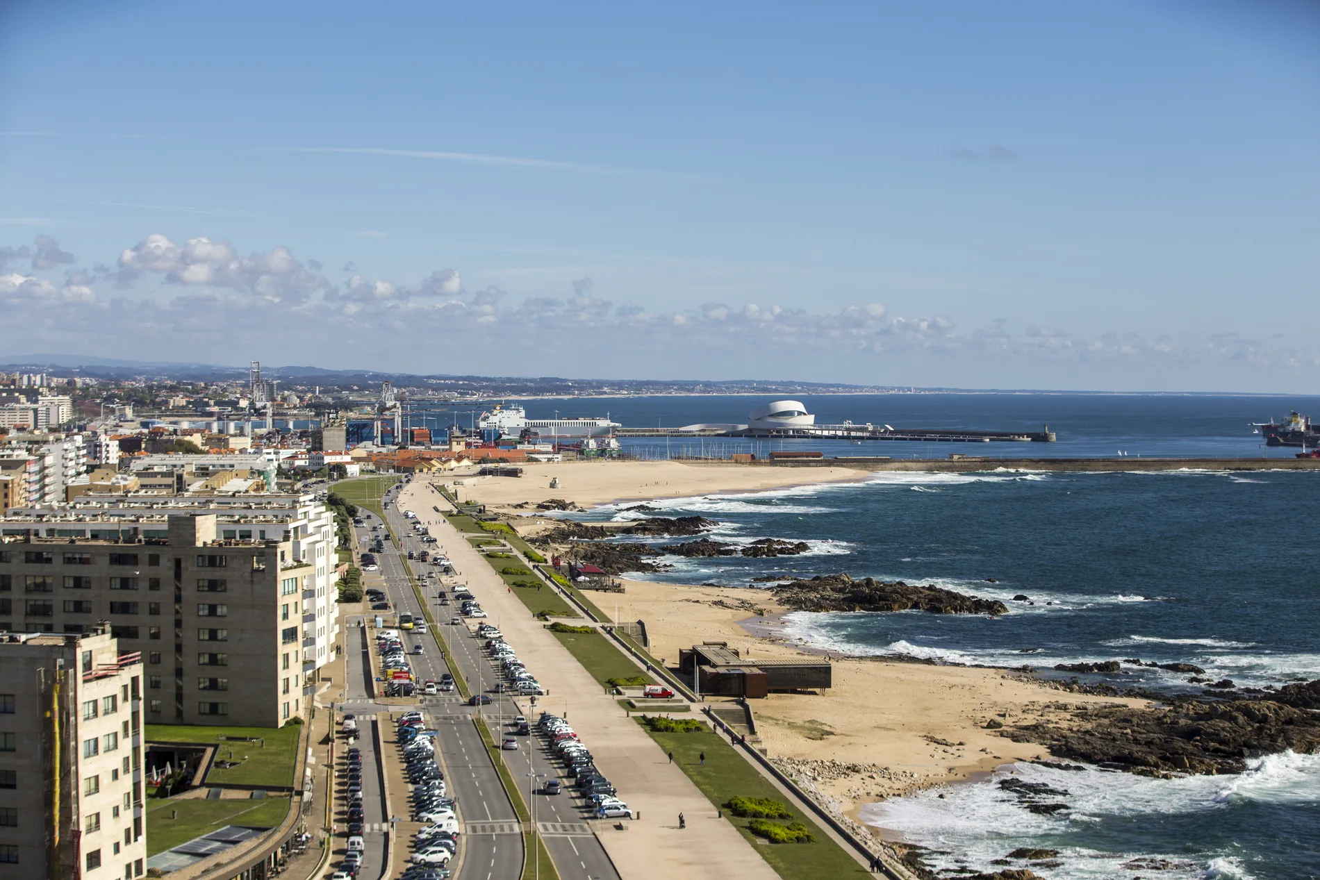 Coastal cityscape with a sandy beach, road, and buildings by the sea under a blue sky.