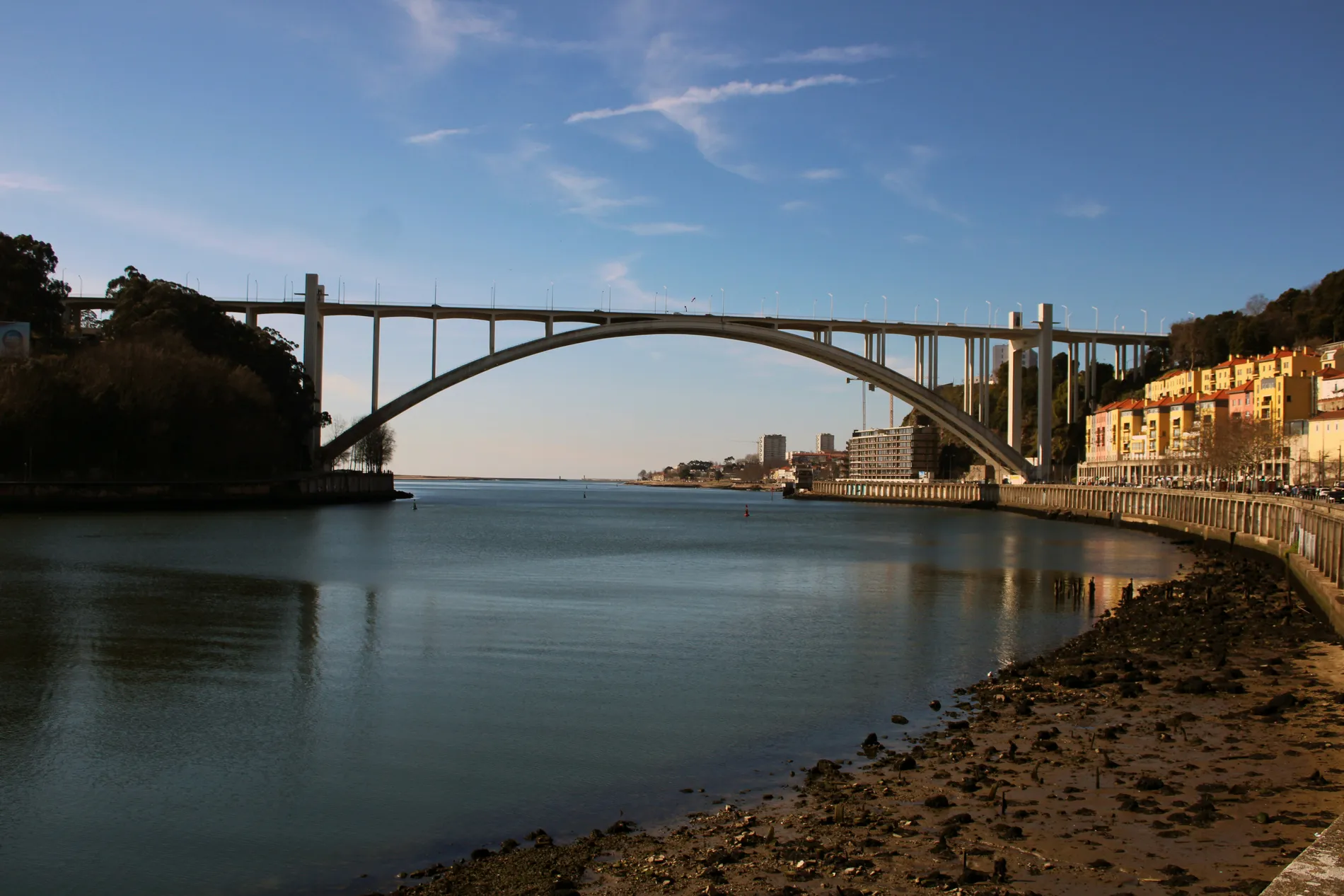 A large arch bridge spans over a calm river, with colorful buildings along the shoreline.