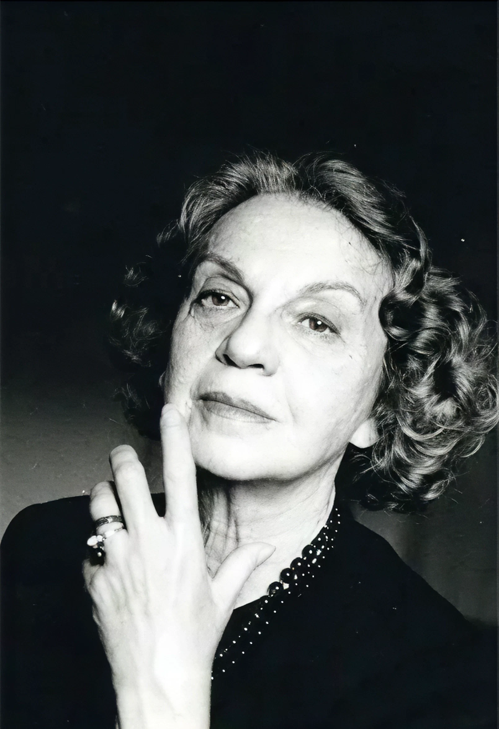 Elderly woman with curly hair, thoughtful expression, hand near face. Black and white image.