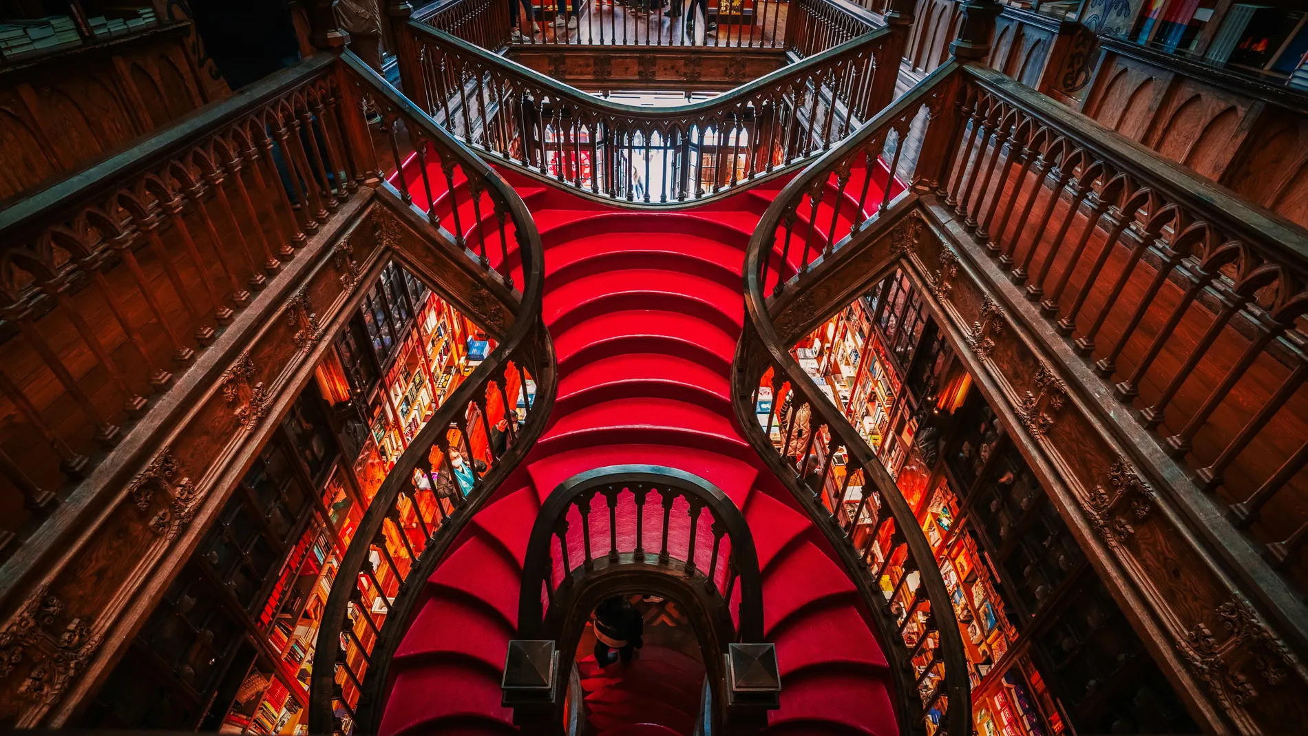 Ornate red spiral staircase with wooden railings in a library setting.