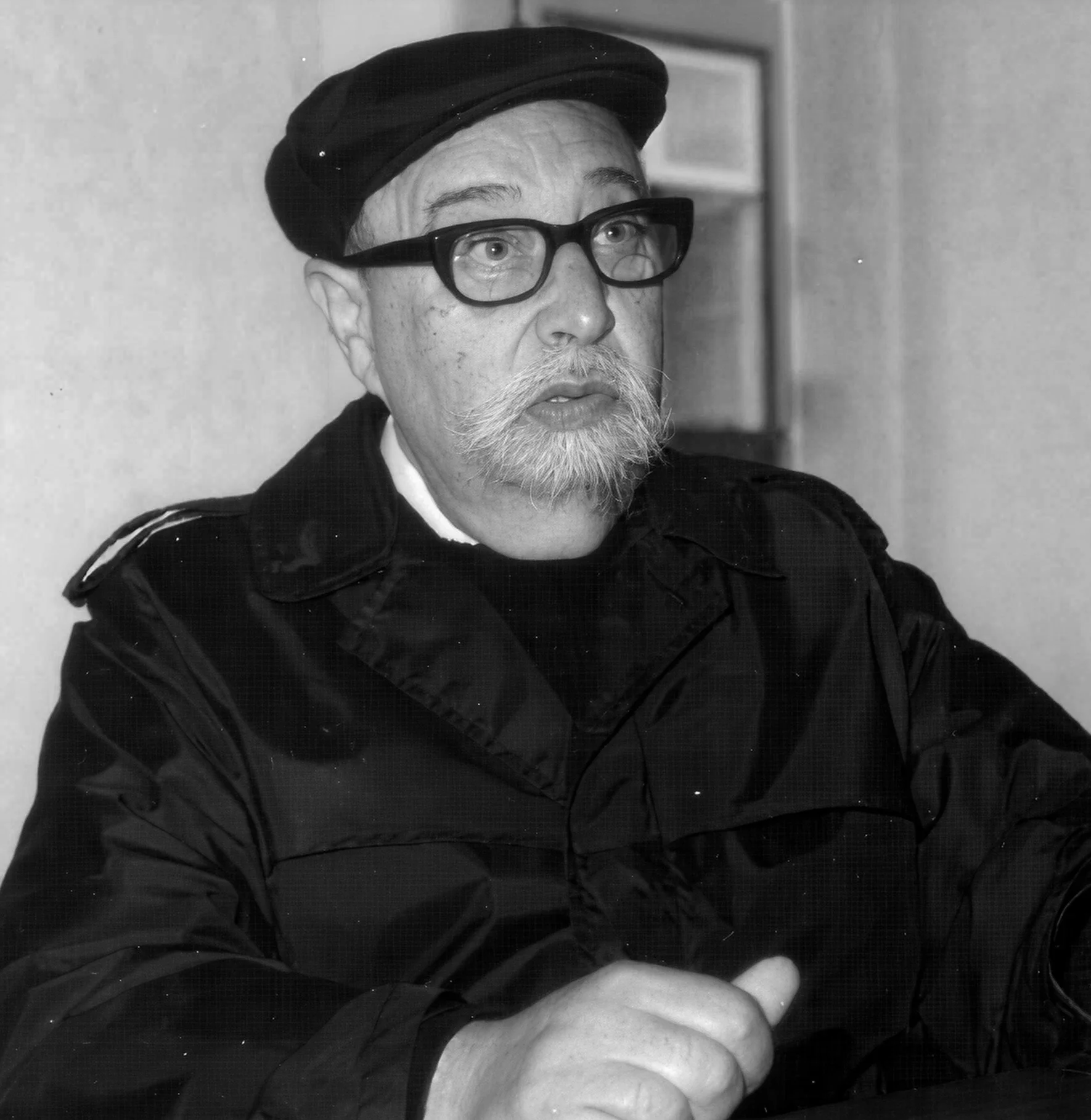 Man with glasses and a beard wearing a cap and jacket, sitting indoors.