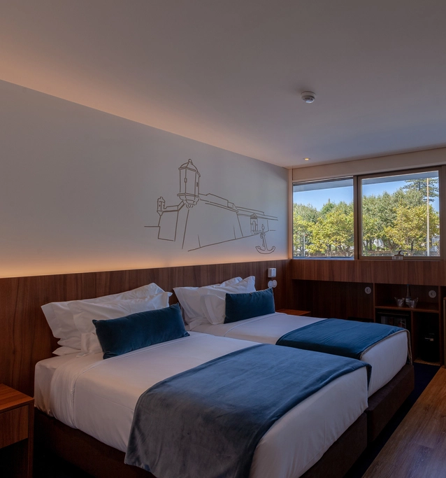 Hotel room with two beds, blue blankets, and a window view of trees. Wall art features a building.