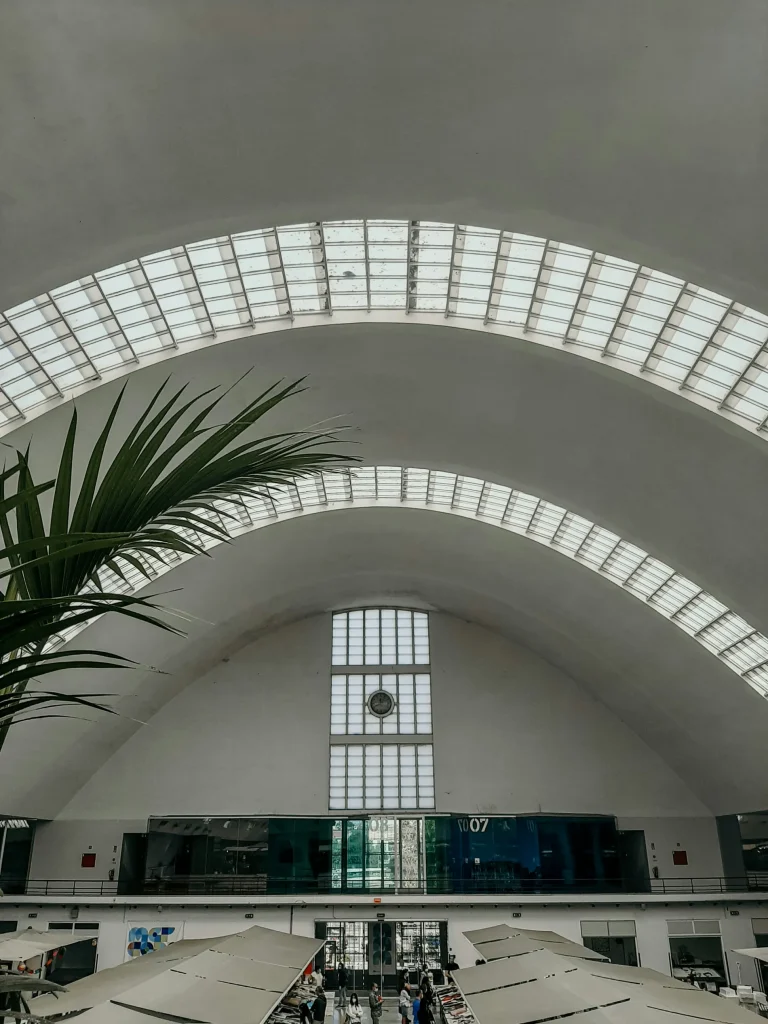 Arched ceiling with skylights, indoor plants, large clock, and people in a spacious hall.