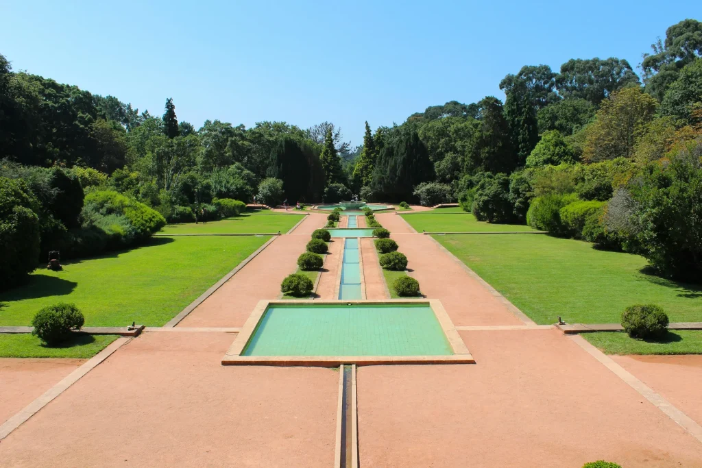 Symmetrical garden with a central fountain, trees, and pathways under a clear blue sky.