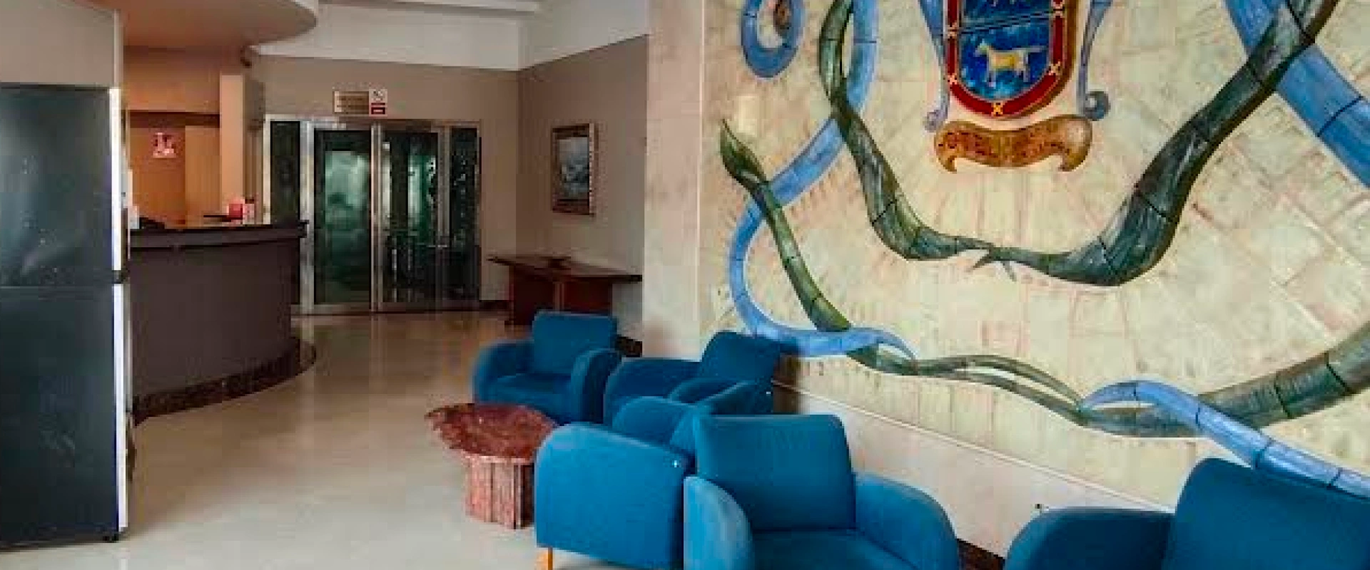 Lobby con sillones azules, mesa de madera y mural decorativo en la pared.