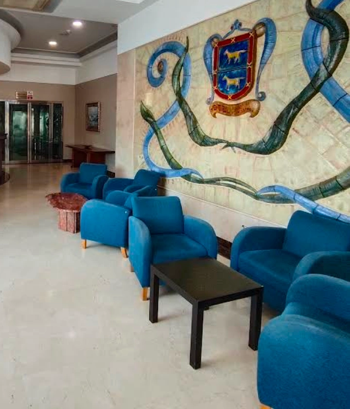 Sala de espera con sillones azules, mesa negra, mural y emblema en la pared.