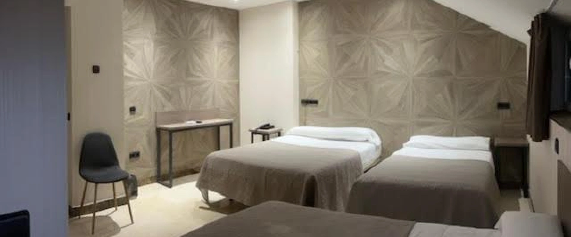 Habitación de hotel con tres camas blancas, escritorio y silla gris, decoración sencilla.