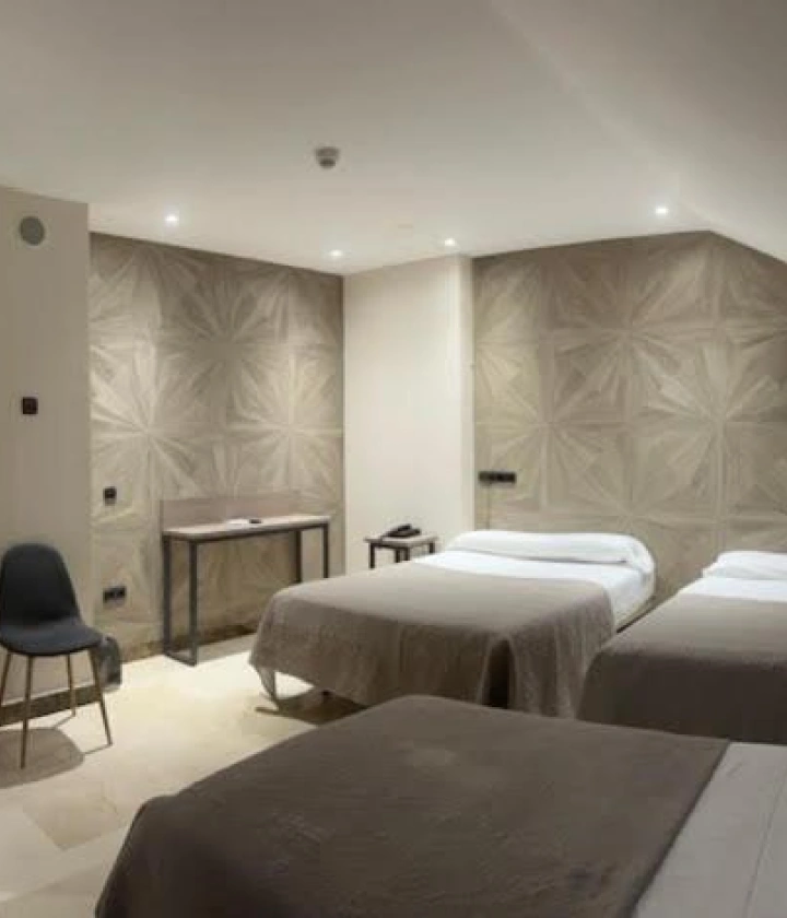 Habitación con dos camas, una silla, escritorio y pared decorativa en tonos beige.