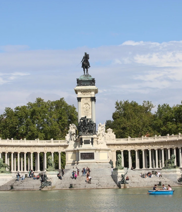 Monumento en un parque con personas y bote en el agua; árboles y cielo despejado al fondo.