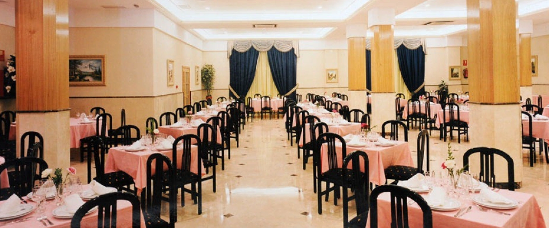 Restaurante elegante con mesas rosadas y sillas negras, columnas y cortinas azules al fondo.
