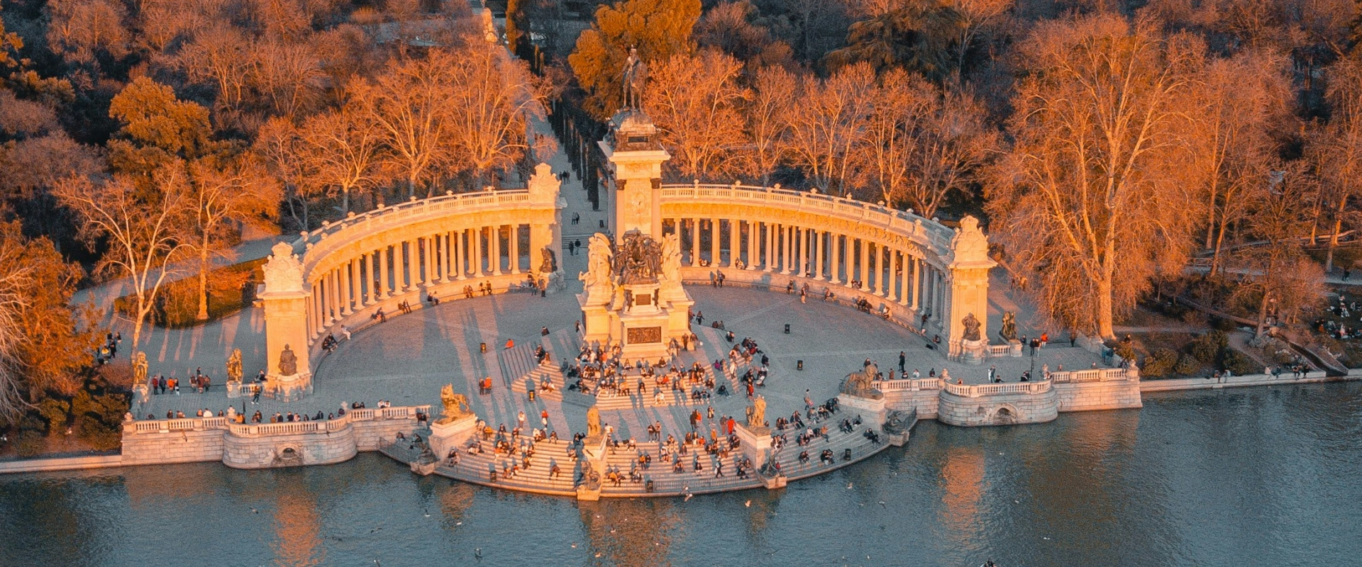 Monumento rodeado de columnas junto a un lago en un parque, con personas alrededor al atardecer.