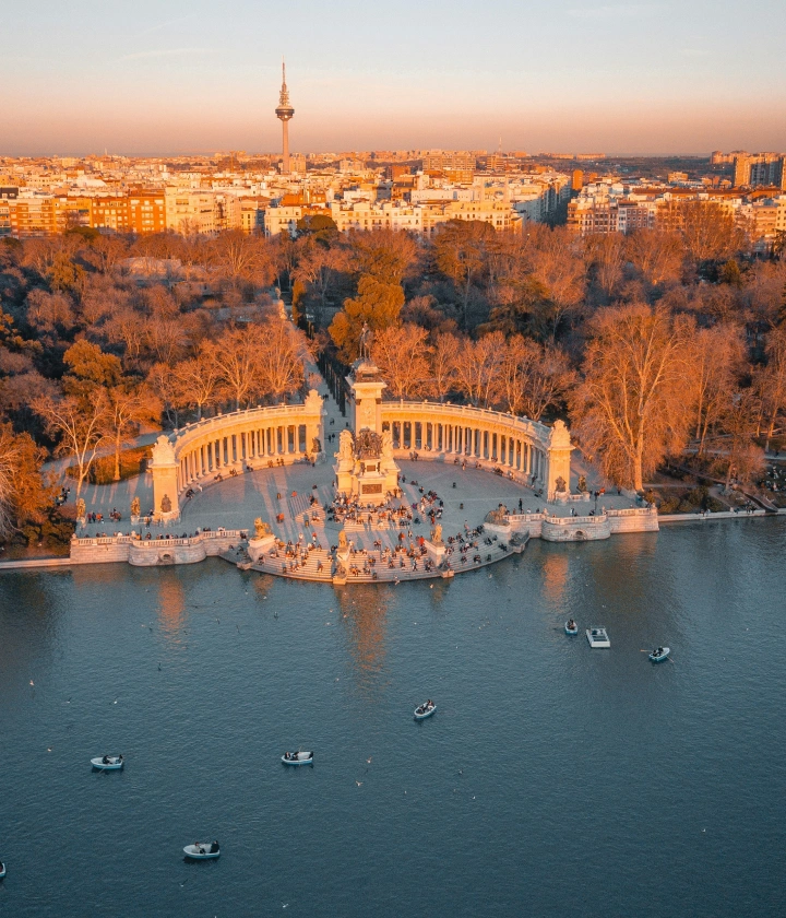 Vista aérea de un monumento en un parque con un lago y botes, edificios al fondo al atardecer.