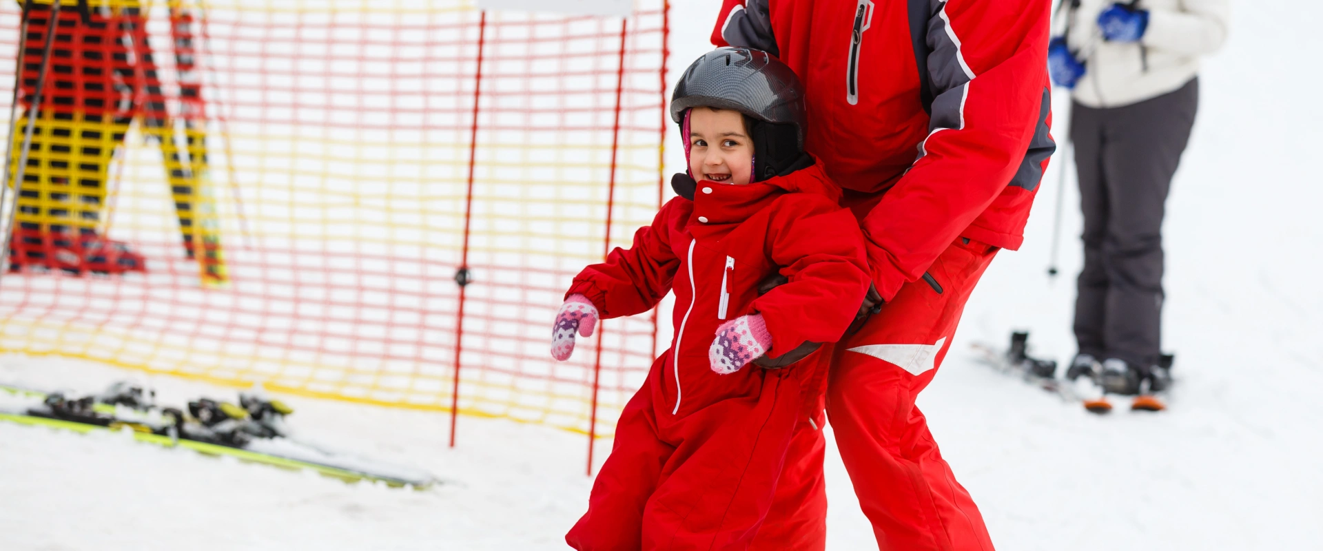 Niño y adulto esquiando con ropa roja y casco en la nieve, cerca de una malla de seguridad.