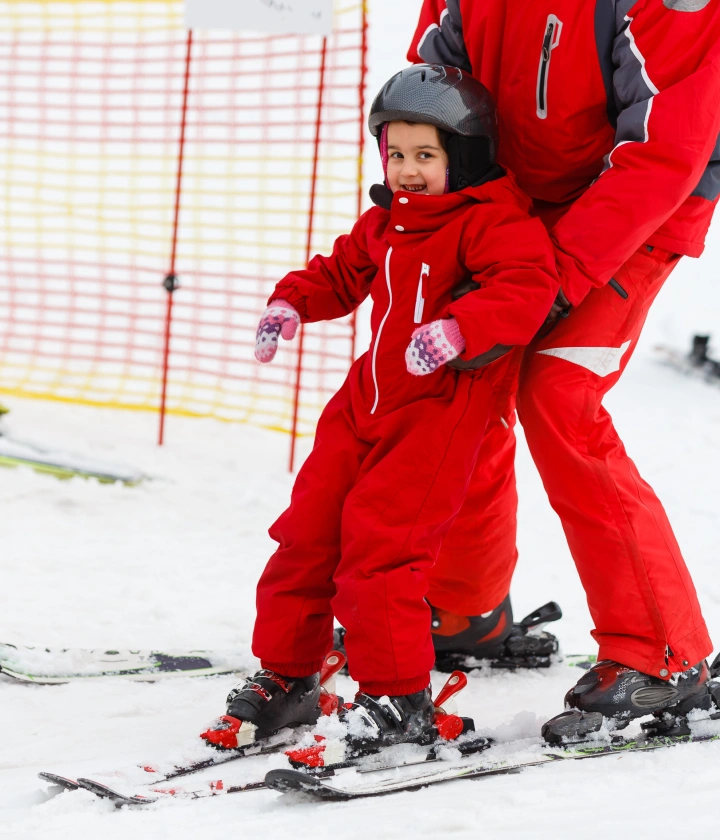 Niño aprendiendo a esquiar con instructor, ambos en trajes rojos, en una pista nevada.