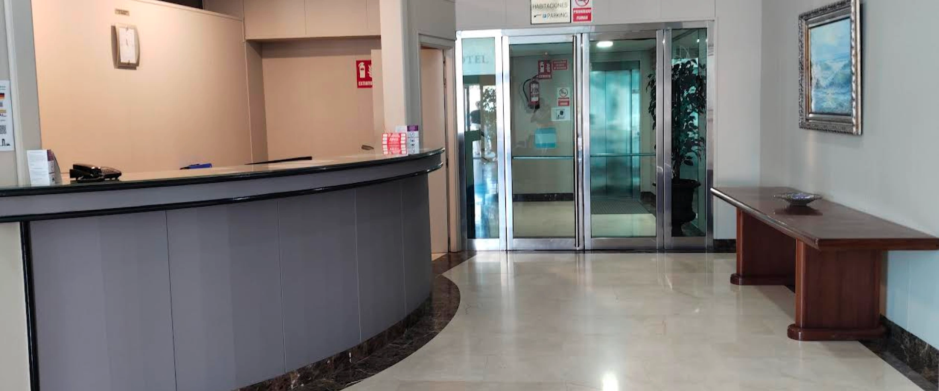 Recepción de hotel con mostrador curvo, puertas de vidrio y mesa de madera.