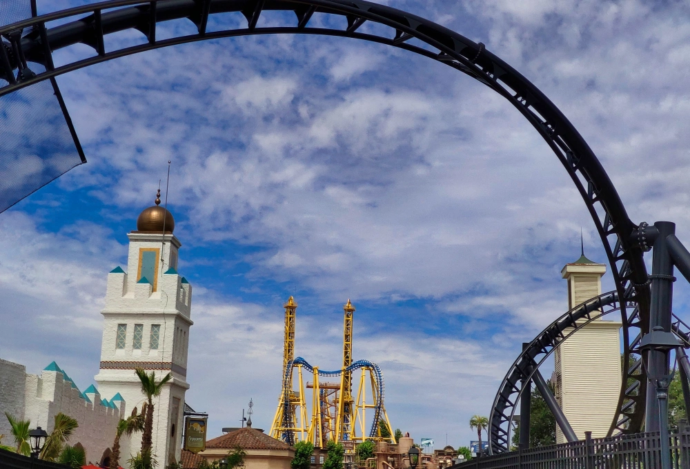 Parque de atracciones con una montaña rusa, cielo nublado y edificios de estilo árabe y occidental.