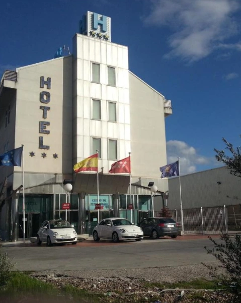 Edificio de hotel de cuatro pisos con banderas y coches estacionados enfrente bajo un cielo azul.