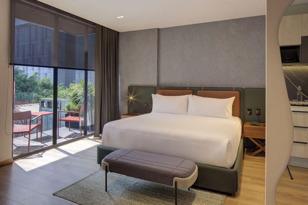 Habitación moderna con cama doble, terraza con sillas y vista al jardín. Decoración en tonos neutros y madera.