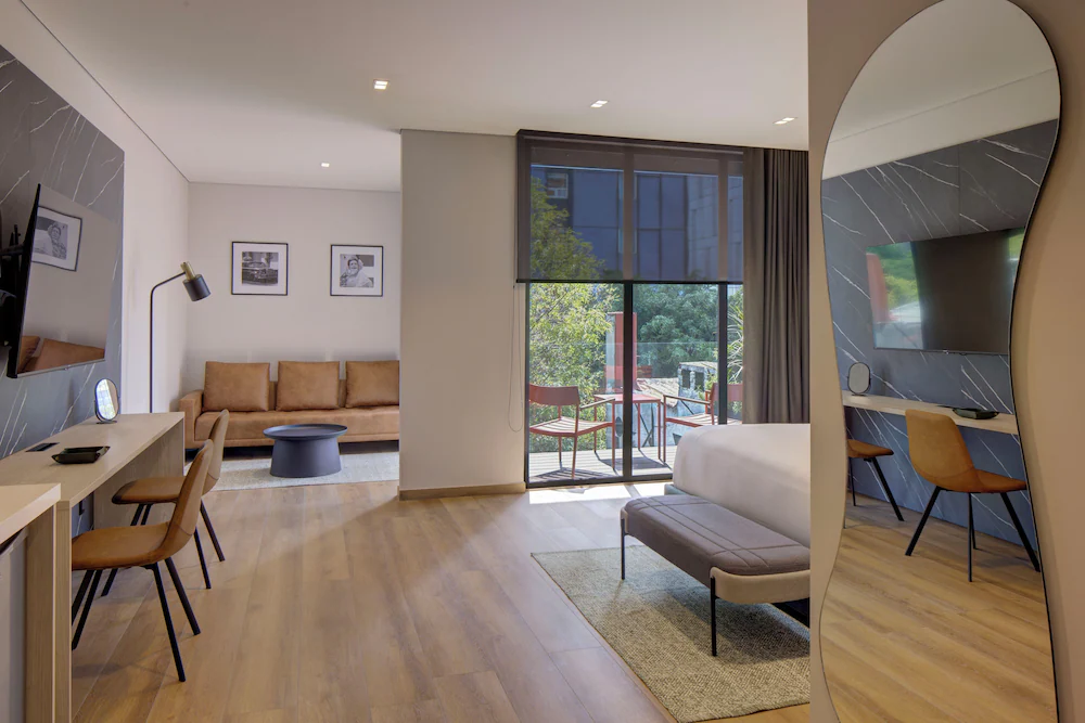Habitación moderna con sofá, escritorio, cama, espejo curvo y balcón con sillas, decorada en tonos neutros.