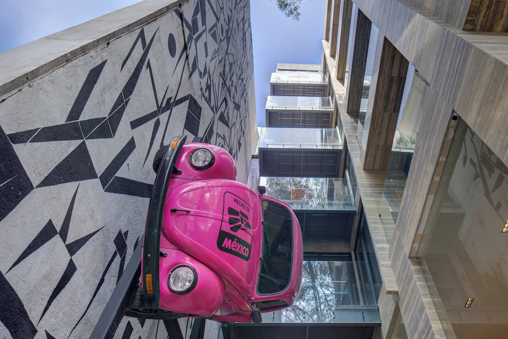 Auto rosa "Vocho" en la pared de un edificio, junto a un mural geométrico blanco y negro en la Ciudad de México.