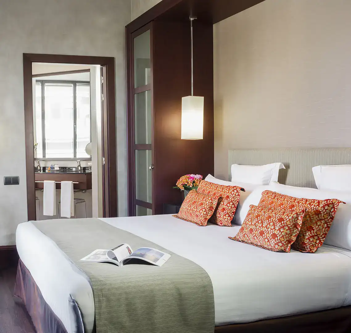 Habitaci&oacute;n de hotel con cama grande, almohadas rojas y puerta al ba&ntilde;o.