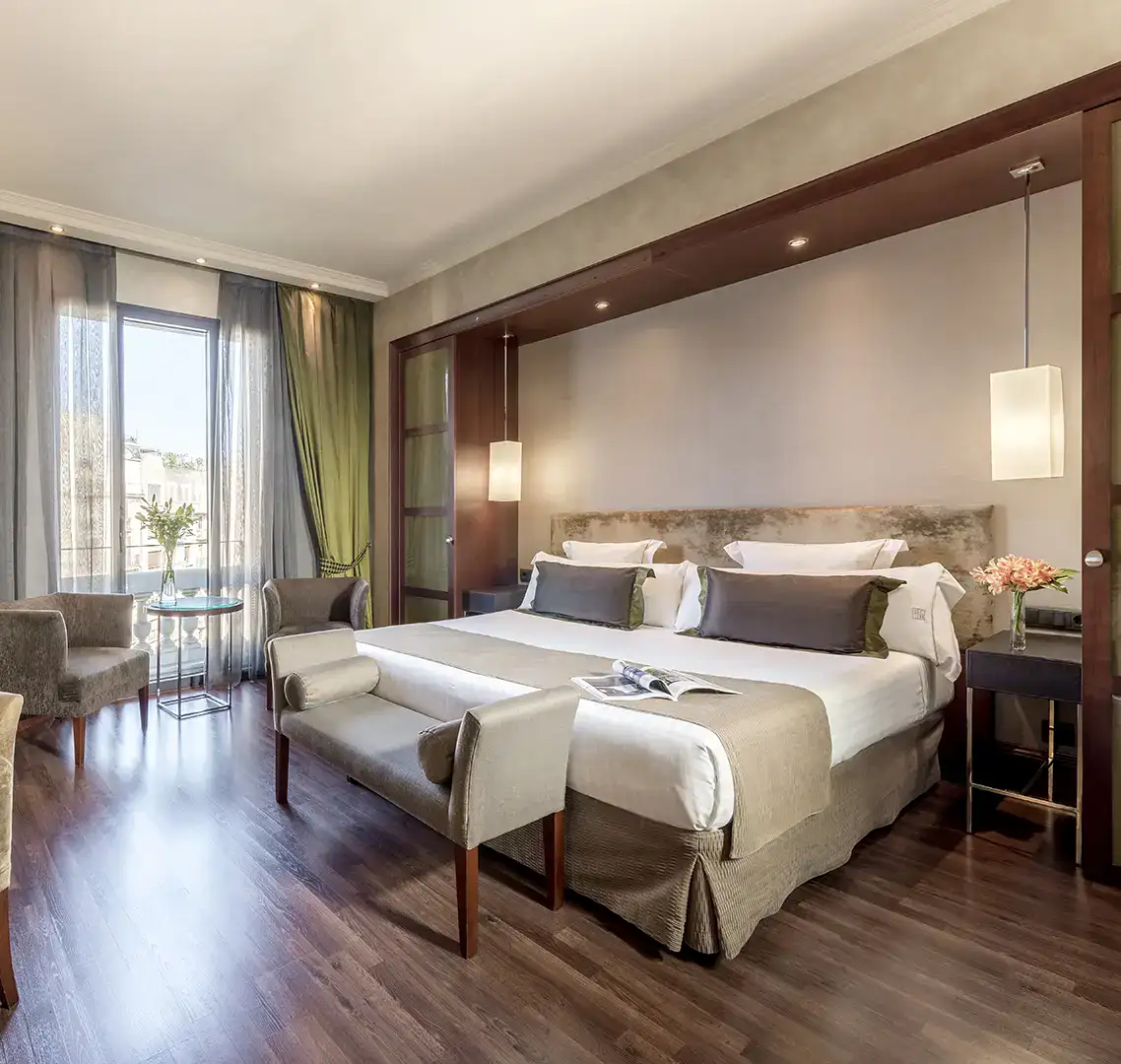 Habitaci&oacute;n de hotel elegante con cama doble, sillones y ventanal.