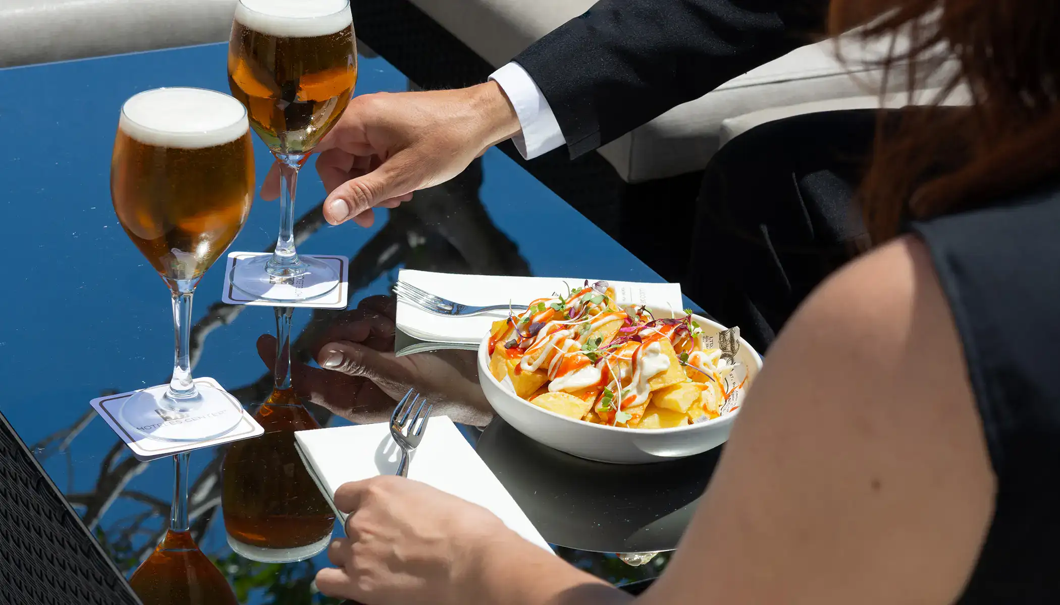 Dos personas compartiendo cervezas y patatas en una mesa de cristal.