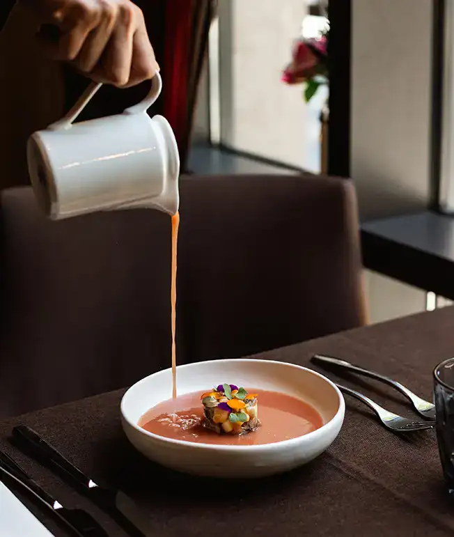 Persona sirviendo sopa color salm&oacute;n sobre plato con comida decorada.