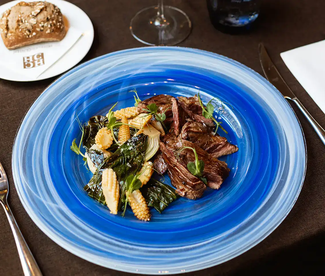 Carne asada con verduras en plato azul junto a pan y cubiertos.