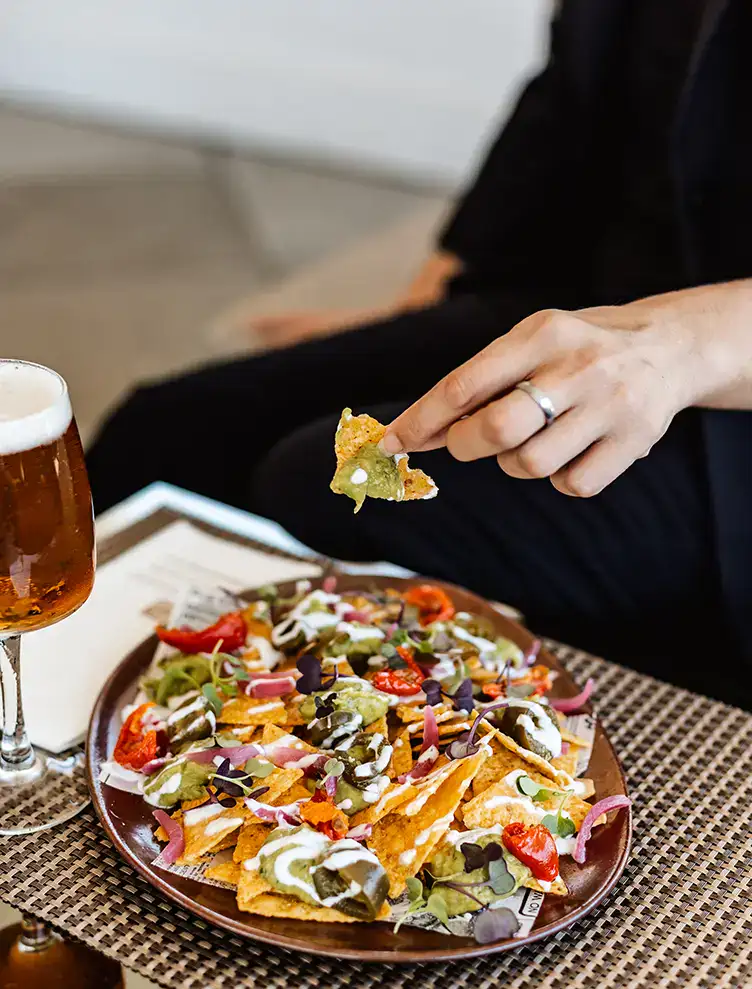 Mano tomando nacho con guacamole, junto a plato de nachos y vaso de cerveza.
