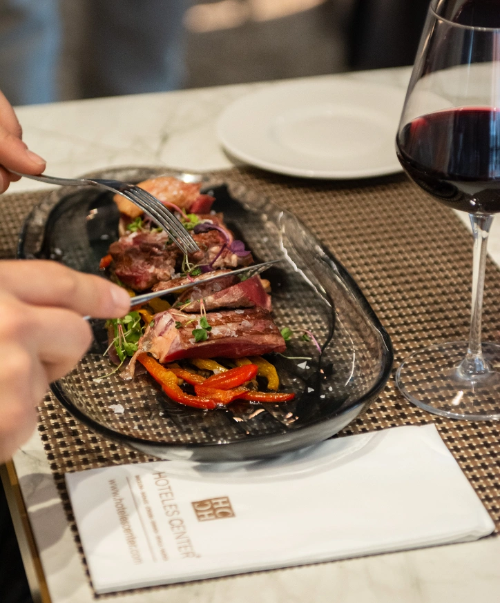 Plato de carne con guarnici&oacute;n y copa de vino tinto en mesa elegante.