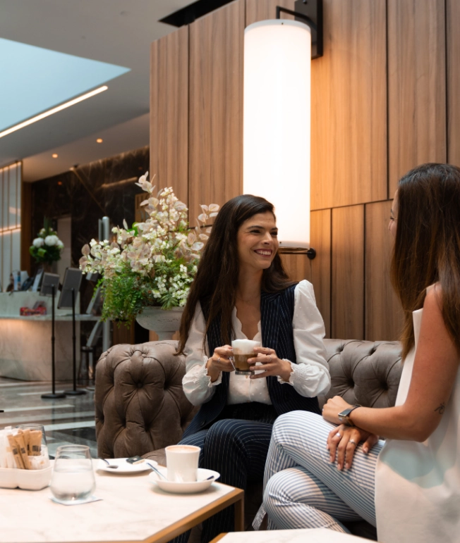 Dos mujeres conversan y beben caf&eacute; en un elegante sal&oacute;n moderno.