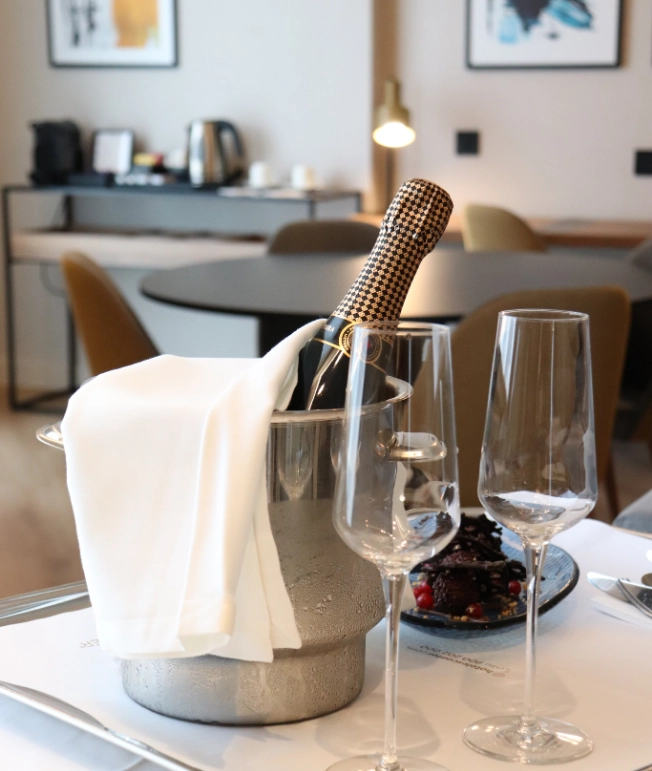 Botella de vino y copas sobre mesa elegante con postre al fondo.