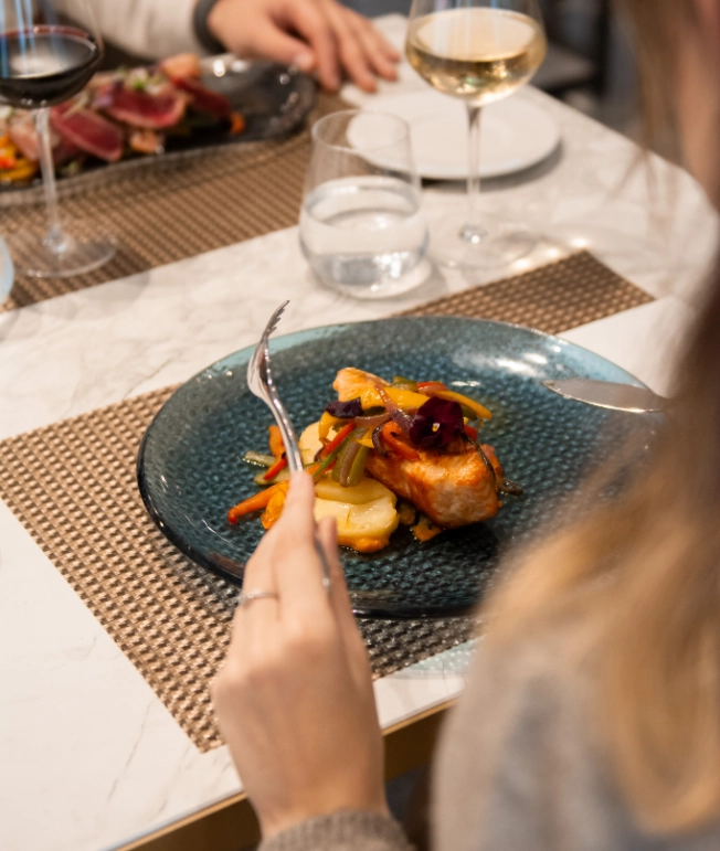 Persona cortando pescado con vegetales en un plato, con copas de vino al lado.