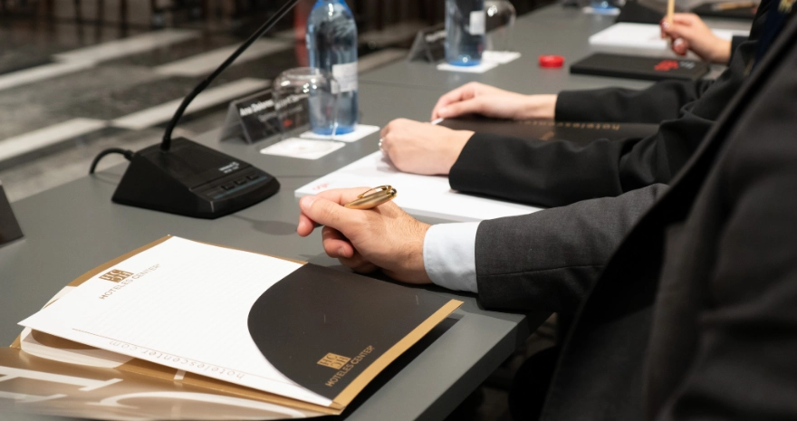 Personas en una mesa de conferencia con libretas y micr&oacute;fonos, listas para tomar notas.