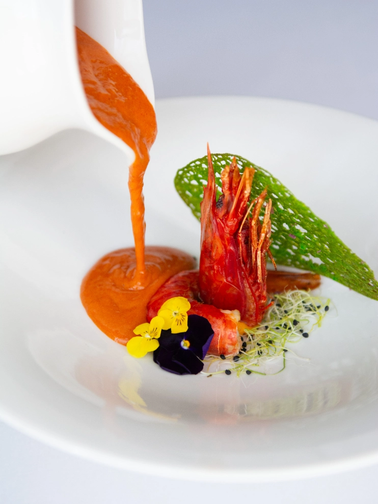 Sopa roja vertida sobre camar&oacute;n y flores comestibles en plato blanco.