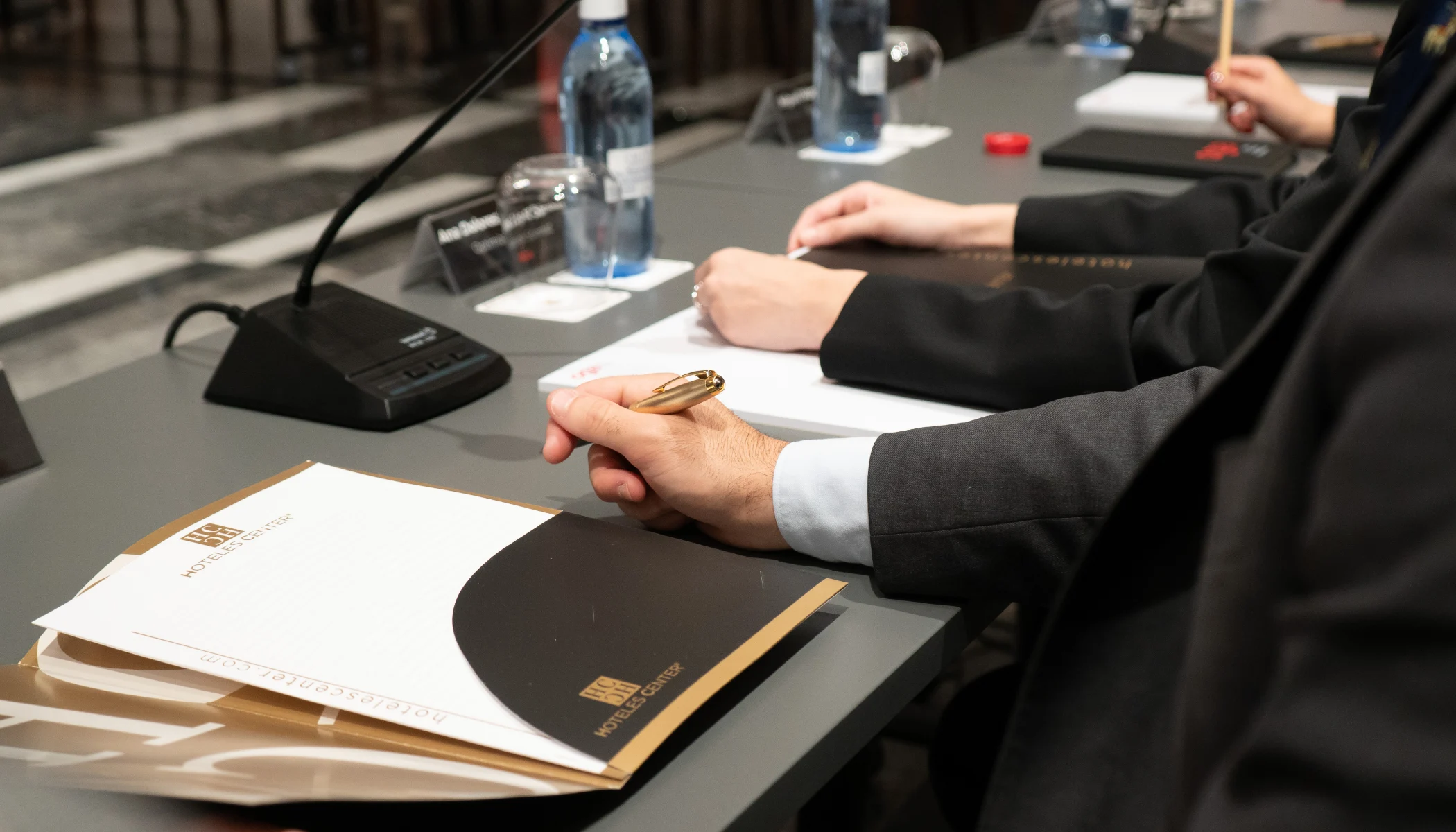 Personas con trajes formales escriben en un escritorio con micr&oacute;fonos.