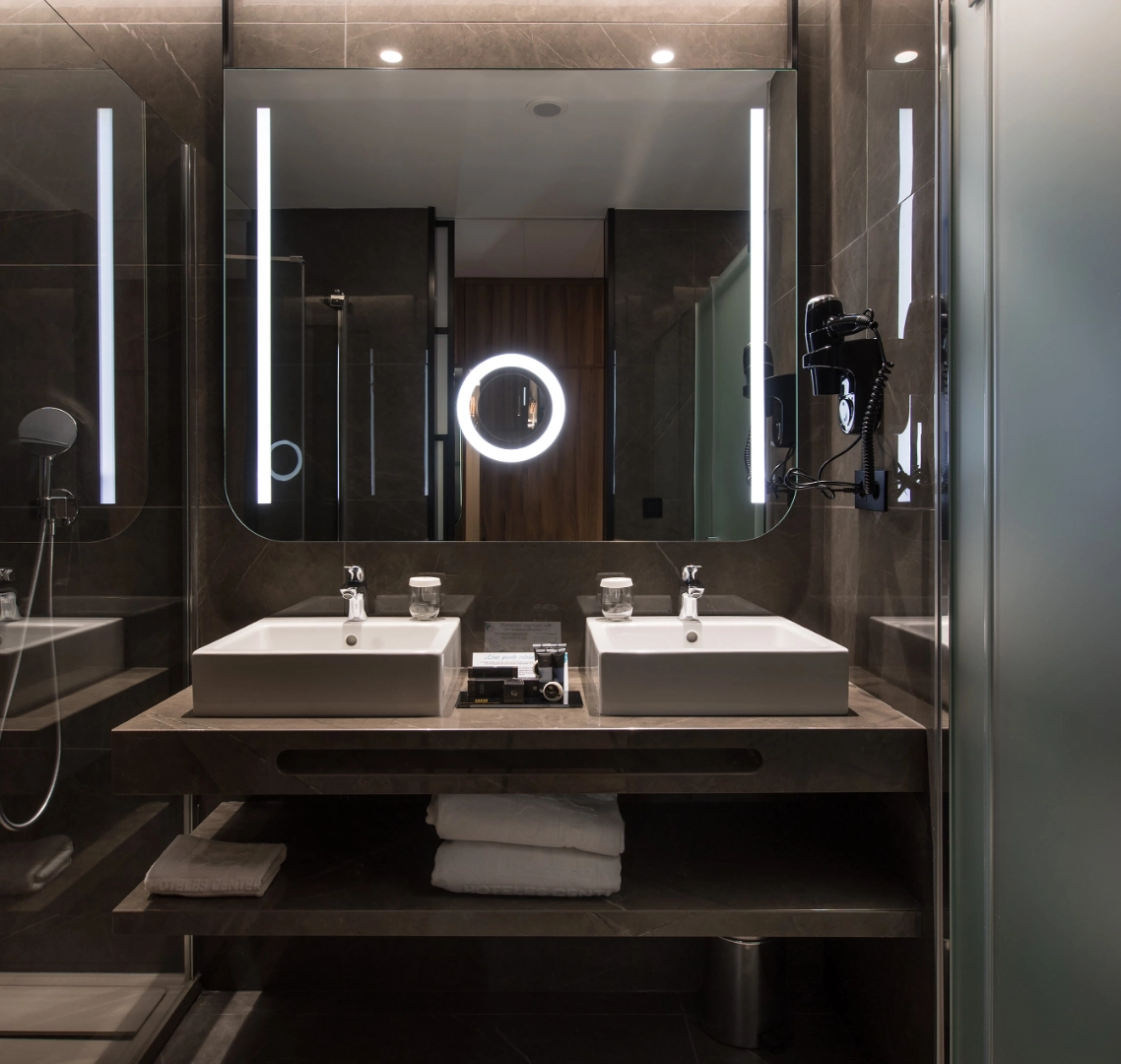 Ba&ntilde;o moderno con lavabo doble, espejo iluminado y toallas dobladas.