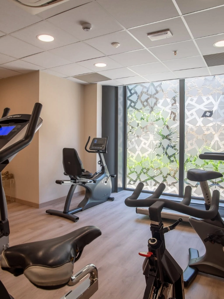 Sala de gimnasio con bicicletas est&aacute;ticas junto a ventanales decorados.