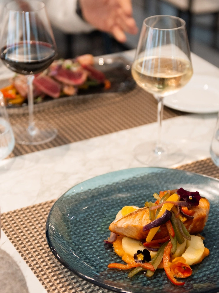 Plato de salm&oacute;n con guarnici&oacute;n de vegetales y dos copas de vino sobre la mesa.