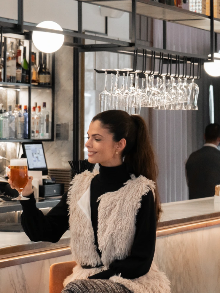 Mujer en un bar sosteniendo un vaso, decorado con copas colgantes.