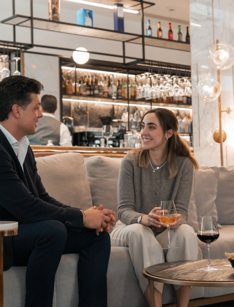Dos personas conversan en un bar mientras toman bebidas en una mesa baja.