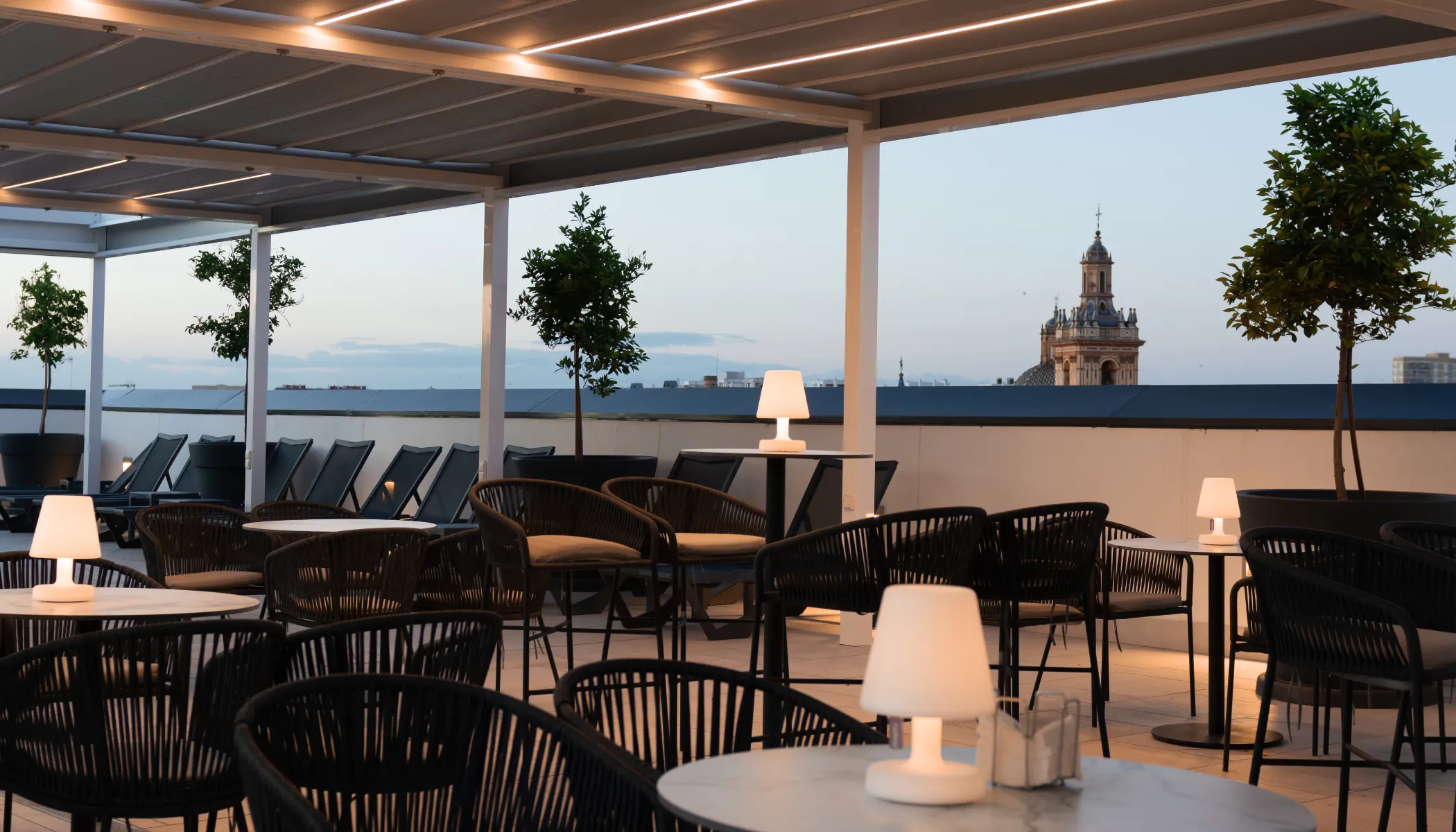 Terraza al atardecer con mesas, sillas y vistas de una torre.