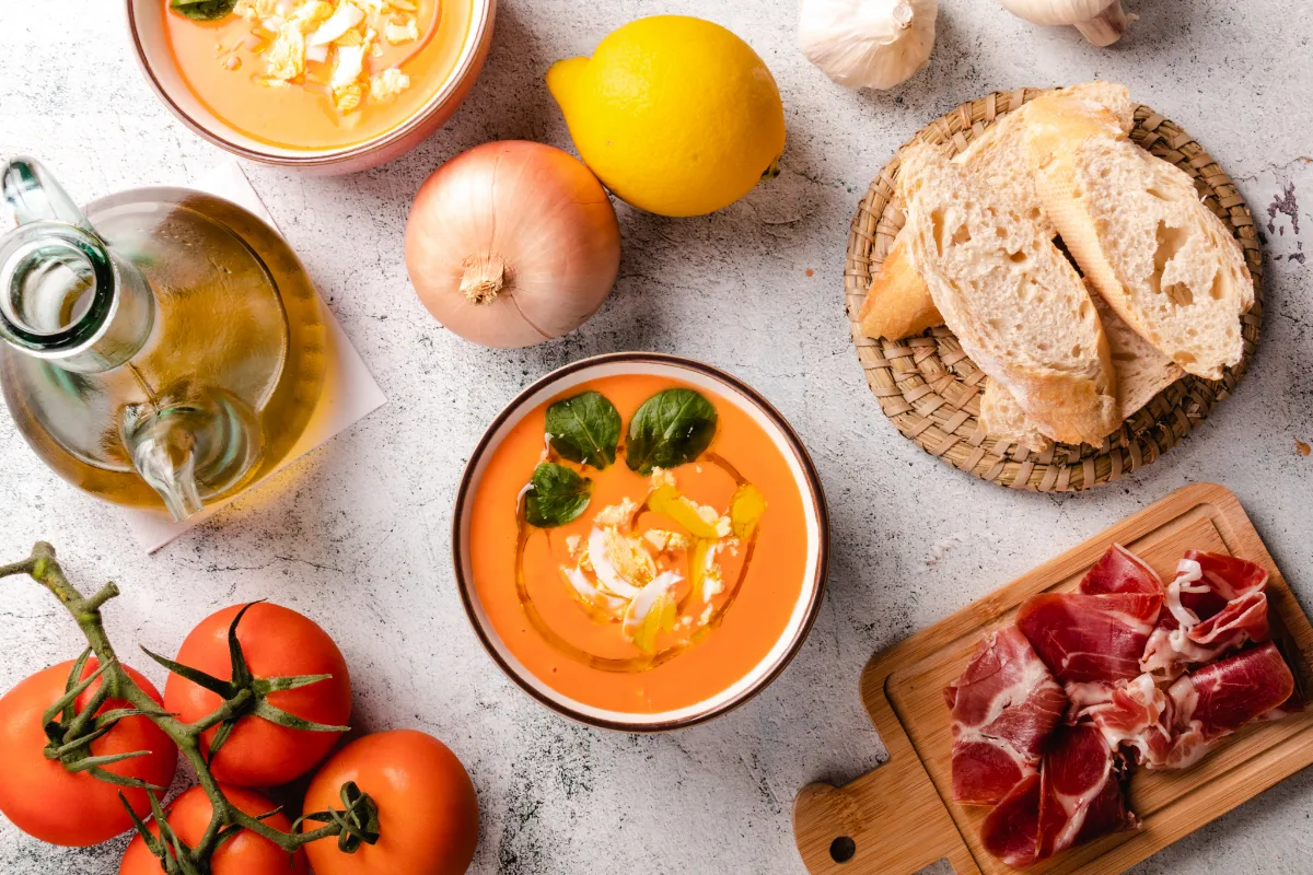 Sopa fr&iacute;a con ingredientes alrededor: pan, jam&oacute;n, tomate, ajo, cebolla y lim&oacute;n.