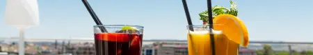 Dos bebidas con pajitas, una roja y otra amarilla, con fondo urbano y cielo azul.