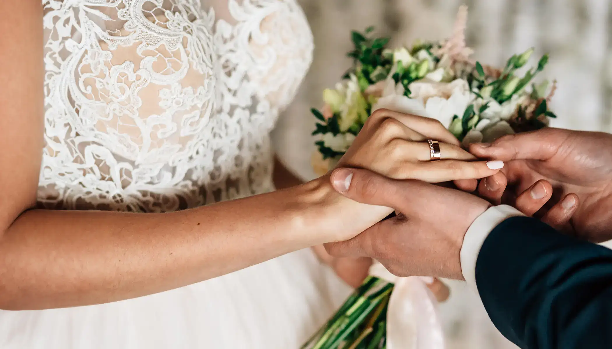 Manos entrelazadas de pareja con vestido de novia y ramo de flores.