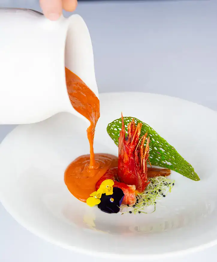 Sopa anaranjada vertida sobre camar&oacute;n y brotes en plato blanco.