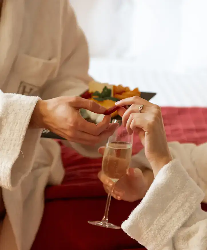 Dos personas en bata compartiendo comida y champ&aacute;n sobre una cama.